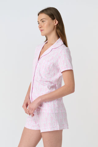 A woman with long, wavy brown hair stands barefoot against a white background, wearing the Lovely Monkey Long Sleeve Polo Pajamas in light pink with a subtle floral pattern—a comfy button-up top and matching pants perfect for relaxing nights.
