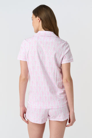 A woman with long, wavy brown hair stands barefoot against a white background, wearing the Lovely Monkey Long Sleeve Polo Pajamas in light pink with a subtle floral pattern—a comfy button-up top and matching pants perfect for relaxing nights.