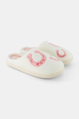 A person stands on a white background wearing Roller Rabbit Lasso Bows Fuzzy Slippers, which feature a cream-colored fuzzy finish, pink horseshoe designs, and durable rubber soles.
