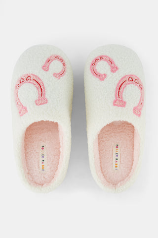 A person stands on a white background wearing Roller Rabbit Lasso Bows Fuzzy Slippers, which feature a cream-colored fuzzy finish, pink horseshoe designs, and durable rubber soles.
