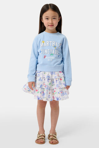 A young girl stands against a plain white background wearing a light blue crewneck sweatshirt that reads “Marthas Vineyard with Roller Rabbit Est. 03,” paired with the Roller Rabbit Kids Monkey On The Vineyard Sami Skirt—a white tiered cotton mini featuring colorful seaside monkey illustrations—and gold sandals.