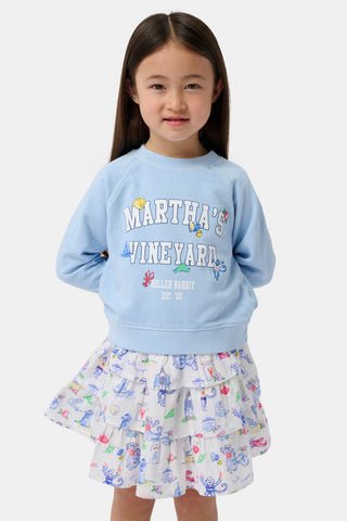 Young girl with long brown hair, wearing a light blue Martha's Vineyard sweatshirt and a white ruffled skirt with colorful doodles, stands facing forward with her hands behind her back and a slight smile, embodying casual seaside style.