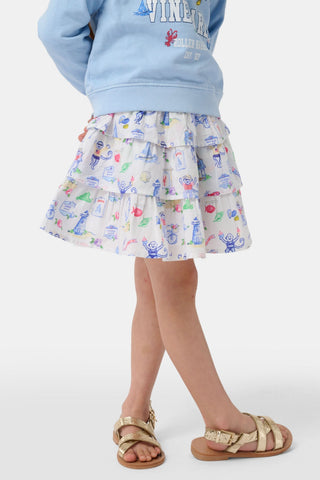 A young girl stands against a plain white background wearing a light blue crewneck sweatshirt that reads “Marthas Vineyard with Roller Rabbit Est. 03,” paired with the Roller Rabbit Kids Monkey On The Vineyard Sami Skirt—a white tiered cotton mini featuring colorful seaside monkey illustrations—and gold sandals.