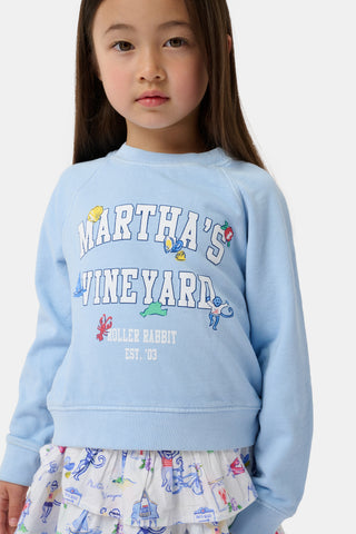 Young girl in a light blue Martha's Vineyard sweatshirt and a white skirt with colorful prints, standing against a plain white background.