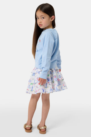 A young girl stands against a plain white background wearing a light blue crewneck sweatshirt that reads “Marthas Vineyard with Roller Rabbit Est. 03,” paired with the Roller Rabbit Kids Monkey On The Vineyard Sami Skirt—a white tiered cotton mini featuring colorful seaside monkey illustrations—and gold sandals.