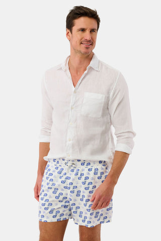 A man stands smiling and looking slightly left against a plain white background, wearing a white linen button-up shirt and Mens Moby Swim Trunk—white, mesh-lined swim shorts with a blue seashell pattern.