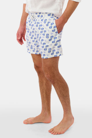 A person models the Mens Moby Swim Trunk—white swim shorts with a blue seashell pattern—paired with a white long-sleeve shirt and stands barefoot against a plain white background.