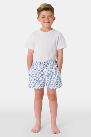 A young boy with light hair wears a plain white T-shirt and Roller Rabbit Boys Moby Swim Trunk. He stands against a white background, one hand in his pocket, smiling slightly and looking to the side.