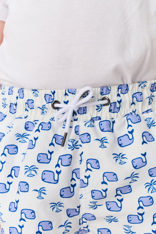 A young boy with light hair wears a plain white T-shirt and Roller Rabbit Boys Moby Swim Trunk. He stands against a white background, one hand in his pocket, smiling slightly and looking to the side.