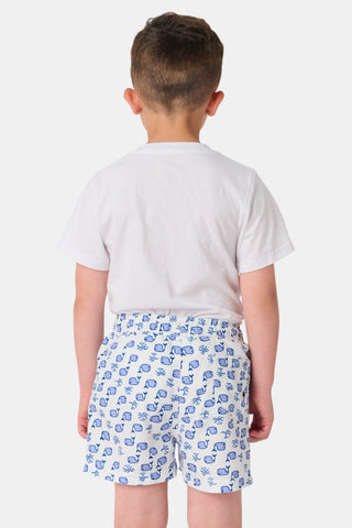 A young boy with light hair wears a plain white T-shirt and Roller Rabbit Boys Moby Swim Trunk. He stands against a white background, one hand in his pocket, smiling slightly and looking to the side.