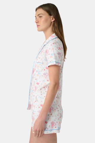 A woman smiles while wearing Roller Rabbit's Monkey Said Yes Polo Pajamas—white short-sleeve top and shorts with colorful, playful prints and blue trim—perfect for bachelorette parties or the newly engaged.