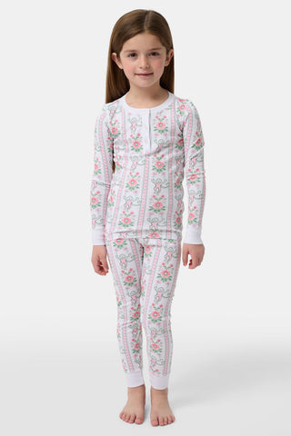 A young girl with long brown hair stands barefoot against a plain background, wearing Roller Rabbit Kids Monkey Block Stripe Pajamas made from soft Pima cotton, featuring playful stripes and monkey motifs.