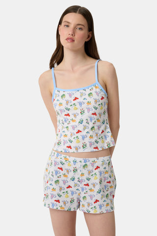 A woman wears the Monkey Fruits D’Ete Clarkson Cami by Roller Rabbit, a sleeveless white cotton-modal cami with light blue trim and a colorful fruit and cartoon print. The matching bottoms are partially visible against a plain white background.