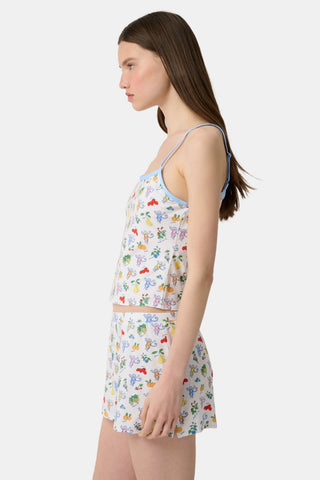A woman wears the Monkey Fruits D’Ete Clarkson Cami by Roller Rabbit, a sleeveless white cotton-modal cami with light blue trim and a colorful fruit and cartoon print. The matching bottoms are partially visible against a plain white background.