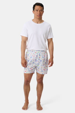 A person wearing a white T-shirt and Roller Rabbit’s Mens Monkey On The Vineyard Boxers, featuring playful and colorful cartoon monkey designs, stands barefoot against a plain white background. The cheerful patterns on the boxers evoke the feel of a summer memento, adding charm to the simple outfit.