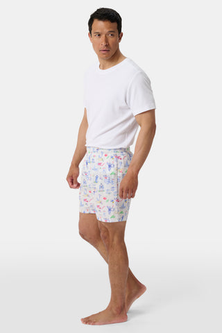 A man stands barefoot wearing a white t-shirt and patterned boxer shorts. He faces slightly to the side, looking off to his left against a minimalist white background.