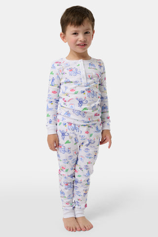 Young child standing barefoot, smiling, wearing white pajamas decorated with colorful animal and vehicle doodle prints. The pajamas feature long sleeves, a buttoned neckline, and fitted cuffs. The background is plain and light-colored.