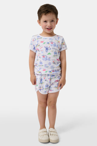 A young child smiles, wearing matching white pajamas with playful cartoon prints and white shoes, standing against a plain white background.