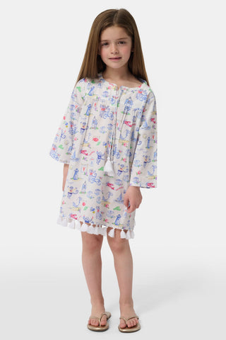 A young girl stands wearing the Roller Rabbit Kids Monkey On The Vineyard Serafina Tunic, a lightweight long-sleeve top styled as a white dress adorned with playful, colorful patterns and finished with ruffle trim at the hem. She has long brown hair and is paired with beige sandals, set against a plain white background.