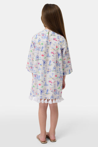 A young girl stands wearing the Roller Rabbit Kids Monkey On The Vineyard Serafina Tunic, a lightweight long-sleeve top styled as a white dress adorned with playful, colorful patterns and finished with ruffle trim at the hem. She has long brown hair and is paired with beige sandals, set against a plain white background.