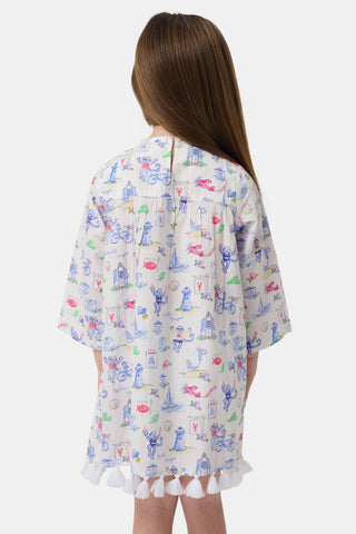 A young girl stands wearing the Roller Rabbit Kids Monkey On The Vineyard Serafina Tunic, a lightweight long-sleeve top styled as a white dress adorned with playful, colorful patterns and finished with ruffle trim at the hem. She has long brown hair and is paired with beige sandals, set against a plain white background.