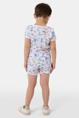 A young child stands barefoot, smiling and facing forward, wearing Roller Rabbit Kids Monkey On The Vineyard Pajamas made of soft Pima cotton with colorful cartoon patterns of animals, vehicles, and buildings. The pajamas feature buttons at the neckline.