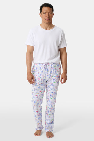 A man stands barefoot wearing a plain white T-shirt and white pajama pants adorned with colorful, playful patterns against a simple light background.