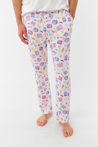 A man stands barefoot against a plain background, wearing the Roller Rabbit Mens Tinned or Twist Milo Set—a Pima cotton T-shirt paired with colorful drawstring waist pants featuring cartoon food patterns.