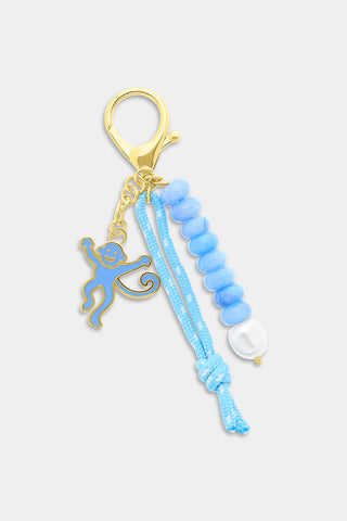 A hand holds two gold keys attached to the Roller Rabbit Monkey Beaded Keychain, featuring blue beads, a smiling monkey charm, a pearl bead, and a blue string, set against a white and blue floral patterned background.