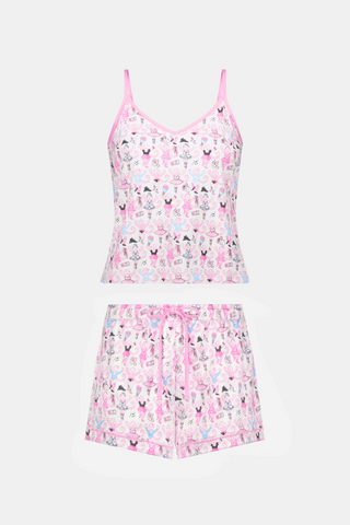 A woman wearing the Roller Rabbit Monkey Pirouette Mischa Set—pink and white cat-print Pima cotton loungewear—and holding a matching bag stands in front of white lockers, smiling slightly at the camera.