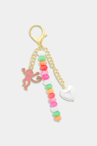 A hand holds gold keys on the Roller Rabbit Monkey Beaded Keychain, featuring colorful beads, a pink smiling monkey charm, and a white pearl charm. The person is wearing a white sweater with pink stripes.