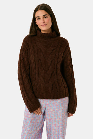 A person wearing a cozy, chocolate brown Naomi Sweater by REVOLUTION KNITS, featuring a cable-knit design and a high turtleneck.