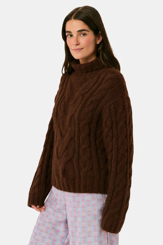 A person wearing a cozy, chocolate brown Naomi Sweater by REVOLUTION KNITS, featuring a cable-knit design and a high turtleneck.