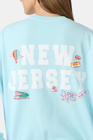 Person wearing a light blue sweatshirt with NEW JERSEY in large white letters on the back, featuring playful illustrations of a hot air balloon, building, airplane, ticket, boat, and lighthouse.