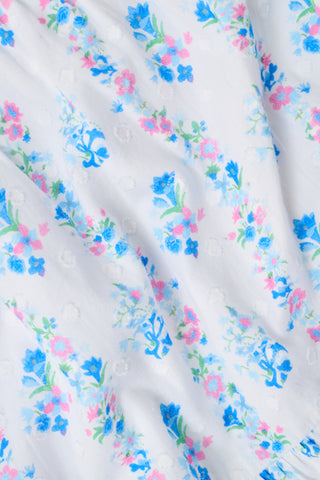 A woman with straight brown hair and a blue beaded necklace wears the Roller Rabbit Naia Floral Stripe Dovie Top—a light blue floral crop top with puff sleeves and a sweetheart neckline—paired with a matching skirt, standing against a plain white background.