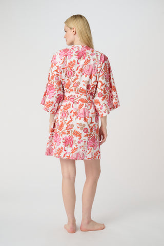 A woman in the Roller Rabbit Amanda Kimono, a pink and white floral cotton robe with wide sleeves and a tie waist, stands against a white background, looking down with a gentle smile.