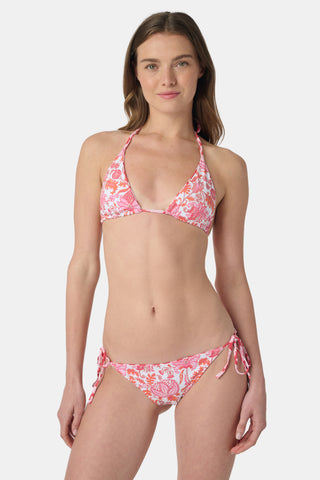 A woman smiles at the beach while holding a straw hat, wearing the Roller Rabbit Amanda/Jemina Serena Bikini Top for stylish sun protection with the ocean and sky behind her.