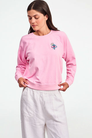 A person wears the Roller Rabbit Palm Beach Monkey Sweatshirt in pastel pink, featuring a colorful blue monkey, sun, and pink flamingo graphic on the left chest; only their upper torso is visible.