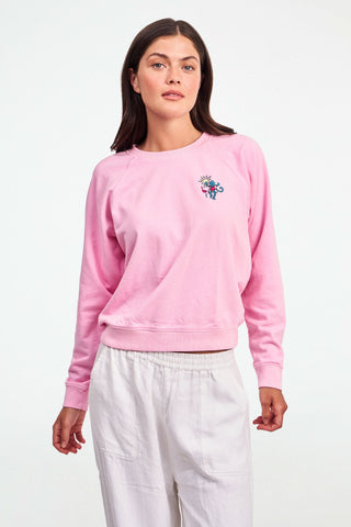 A person wears the Roller Rabbit Palm Beach Monkey Sweatshirt in pastel pink, featuring a colorful blue monkey, sun, and pink flamingo graphic on the left chest; only their upper torso is visible.