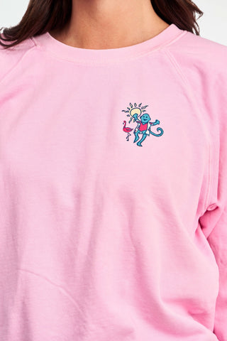 A person wears the Roller Rabbit Palm Beach Monkey Sweatshirt in pastel pink, featuring a colorful blue monkey, sun, and pink flamingo graphic on the left chest; only their upper torso is visible.