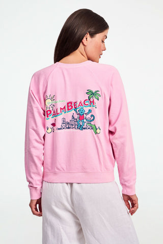 A person wears the Roller Rabbit Palm Beach Monkey Sweatshirt in pastel pink, featuring a colorful blue monkey, sun, and pink flamingo graphic on the left chest; only their upper torso is visible.