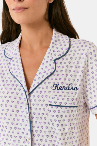 A woman wears the Roller Rabbit Hearts Polo Pajamas, made from 100 cotton with a white base and small heart print. The set features a short-sleeve, button-up collared top with chest pocket and matching shorts. She stands facing forward against a plain background.