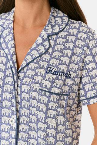 A woman wears the Hathi Polo Pajamas by Roller Rabbit, a premium cotton loungewear set with blue and white elephant patterns, including a collared button-up top and shorts, against a plain light background.