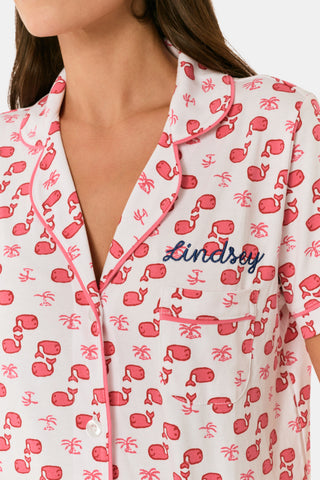 A woman wears Roller Rabbit's Moby Polo Pajamas—a women's short sleeve set with a white base and pink-red cartoon fruit prints, button-up top, and shorts. She stands against a plain light background, smiling gently.