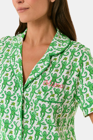 A woman wears Roller Rabbit's Monkey Polo Pajamas, a matching green and white monkey-patterned set with a button-up top and shorts. She touches her hair and smiles softly, looking slightly to the side in her stylish sleepwear.