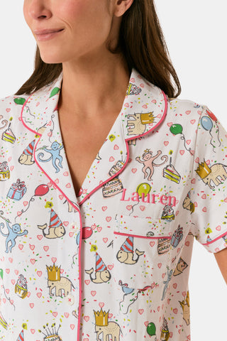 A woman smiles in the Roller Rabbit Birthday Bonanza Polo Pajamas—a women’s short set made from soft Pima cotton with colorful balloon, cake, and animal prints on white.
