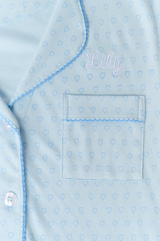 A woman wears Roller Rabbit’s Hearts Tonal Wyatt Polo Pajamas—light blue short-sleeve button-up top with collar and chest pocket, and matching ruffle-hem shorts—standing against a plain white background.
