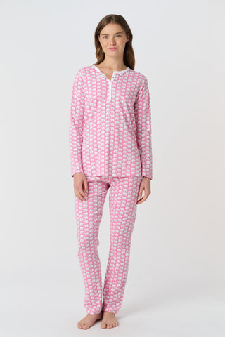 A woman stands barefoot against a plain background, wearing Roller Rabbit's Hathi Pajamas—a matching pink and white set with long sleeves and pants. She faces forward, smiling softly.