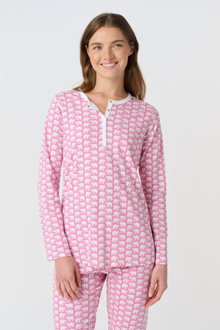 A woman stands barefoot against a plain background, wearing Roller Rabbit's Hathi Pajamas—a matching pink and white set with long sleeves and pants. She faces forward, smiling softly.