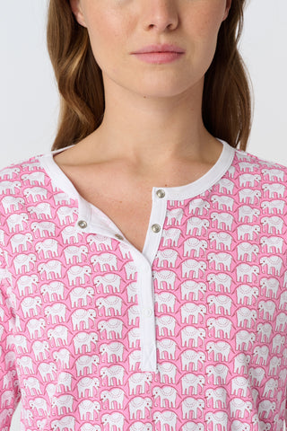 A woman stands barefoot against a plain background, wearing Roller Rabbit's Hathi Pajamas—a matching pink and white set with long sleeves and pants. She faces forward, smiling softly.